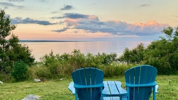 Deux chaises Adirondack bleues, placées face à un plan d’eau, avec un coucher de soleil en arrière-plan.