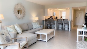 Ein Wohnzimmer mit Sofa, Couchtisch und Sesseln, ein Essbereich mit Bar sowie eine Küche im Hintergrund.