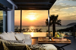 A modern living room with a large window overlooking a sunset over the sea, a wooden coffee table with fruits and a bottle, and a cozy sofa with cushions. - Villa Selenia (Saronikos)