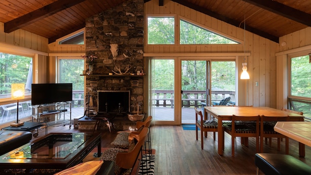 L’intérieur chaleureux d’un chalet avec une cheminée en pierre, des meubles en bois et de grandes fenêtres donnant sur la forêt environnante.