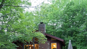 Une cabane rustique dotée d’une terrasse en bois, d’une cheminée en briques et d’une porte en verre donnant sur l’intérieur.