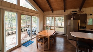L'intérieur d'un chalet en bois comprend une table à manger, des chaises, un coin cuisine et de grandes fenêtres qui offrent une vue sur l'extérieur.