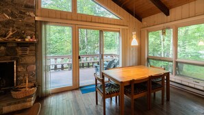 Une table à manger en bois, des chaises, une cheminée en pierre et de grandes fenêtres donnant sur une terrasse.
