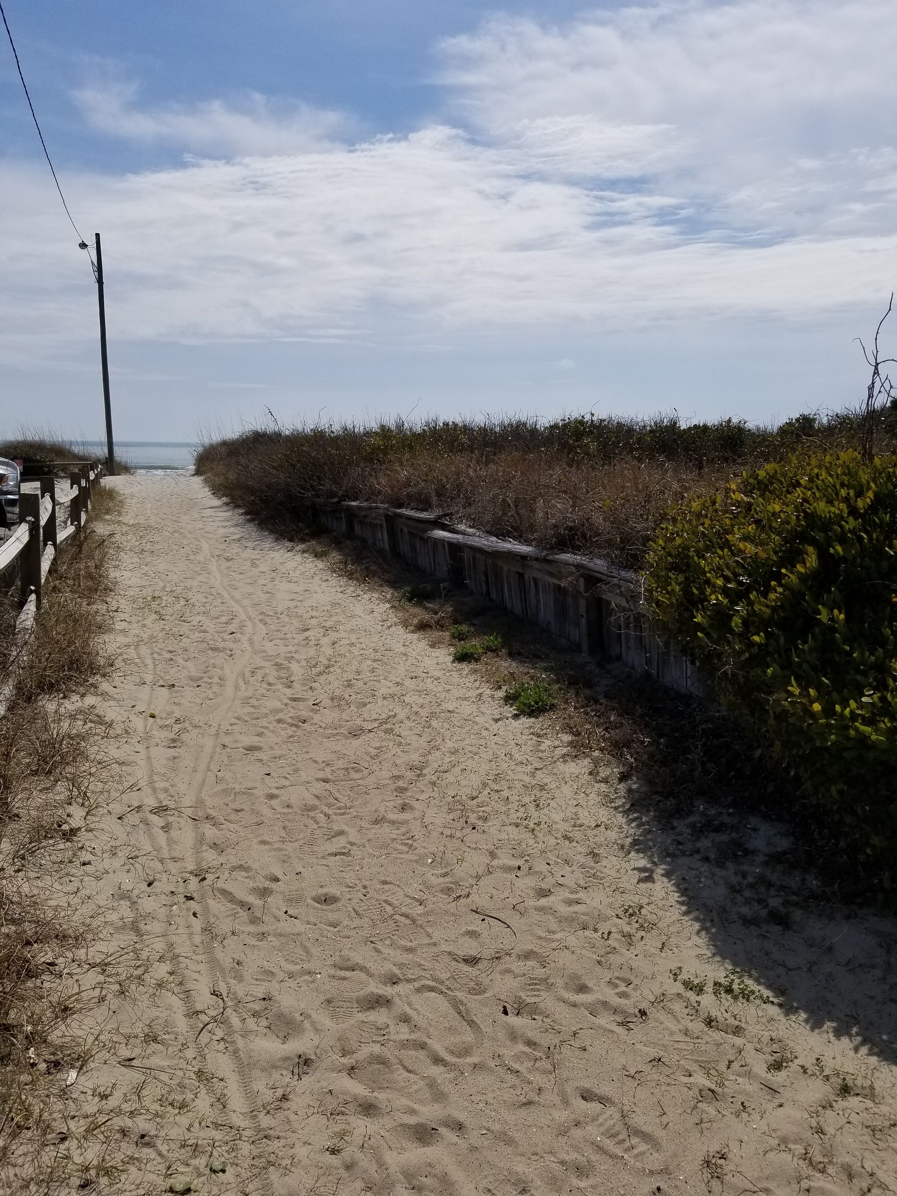 A sandy path leading to a beach with a wooden fence on one side and dense vegetation on the other.