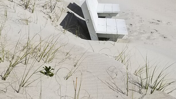 Ein sandiger Strand mit einer weißen Konstruktion, möglicherweise eine Bank oder Sitzgelegenheit, umgeben von hohem Gras und einem Holzzaun.