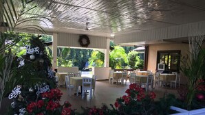 Un patio techado al aire libre con mesas y sillas blancas, un árbol de Navidad y vistas al follaje.