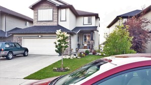 A two-story house with a garage, a driveway, and a small front yard with a tree and a flower bed. - Bright, Welcoming Mountain Escape! Near Downtown! (Calgary)