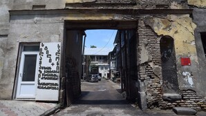 A narrow street with a building entrance featuring a large sign with text, a car parked in front, and a partially visible door. - Wine underground style appartment in the oldest town (Tbilisi)
