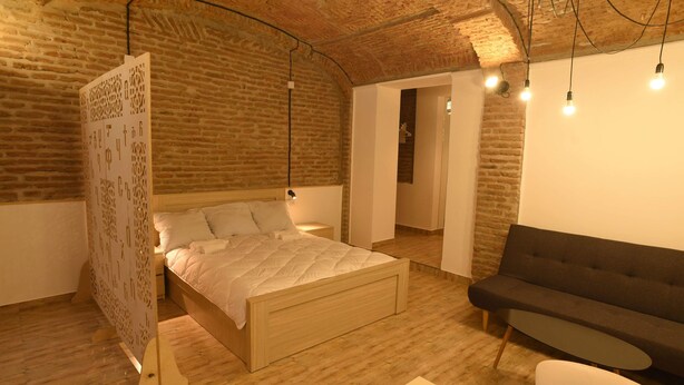 A bedroom with a wooden bed, a dark sofa, and a brick wall. - Wine underground style appartment in the oldest town (Tbilisi)