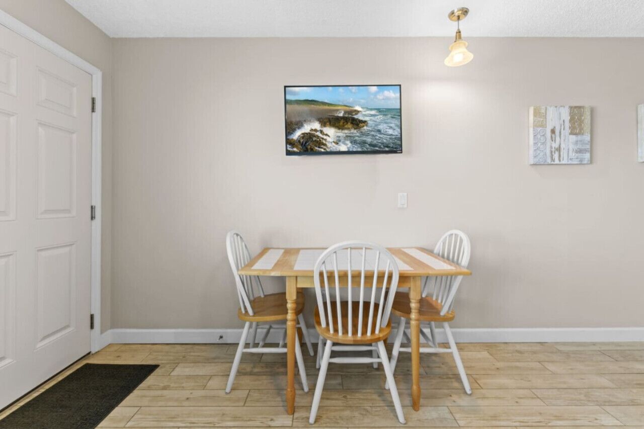 Un comedor con mesa de madera y sillas blancas, un cuadro enmarco con escena costera, y una lámpara.