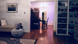 Un salon comprenant un canapé, un tapis à motifs, un tabouret rond en osier, un coin repas avec une table et des chaises, et une bibliothèque remplie de livres et d’objets décoratifs.