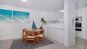 Una cocina moderna con armarios blancos, un microondas y un horno. Hay una zona de comedor con una mesa y sillas de madera, una decoración de un pequeño velero y una planta.