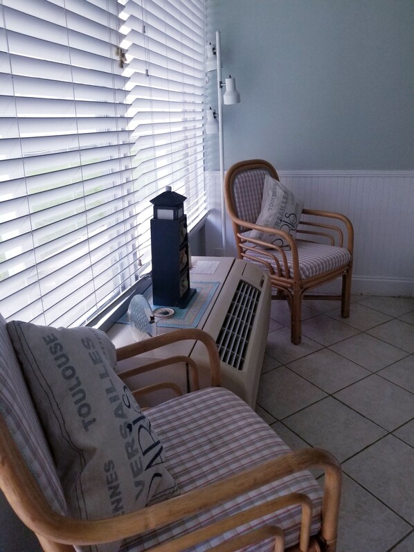 A room with two wicker chairs, one with a striped cushion and the other with a pillow labeled "TOULOUSE". There's a small table with a lamp and a decorative item.