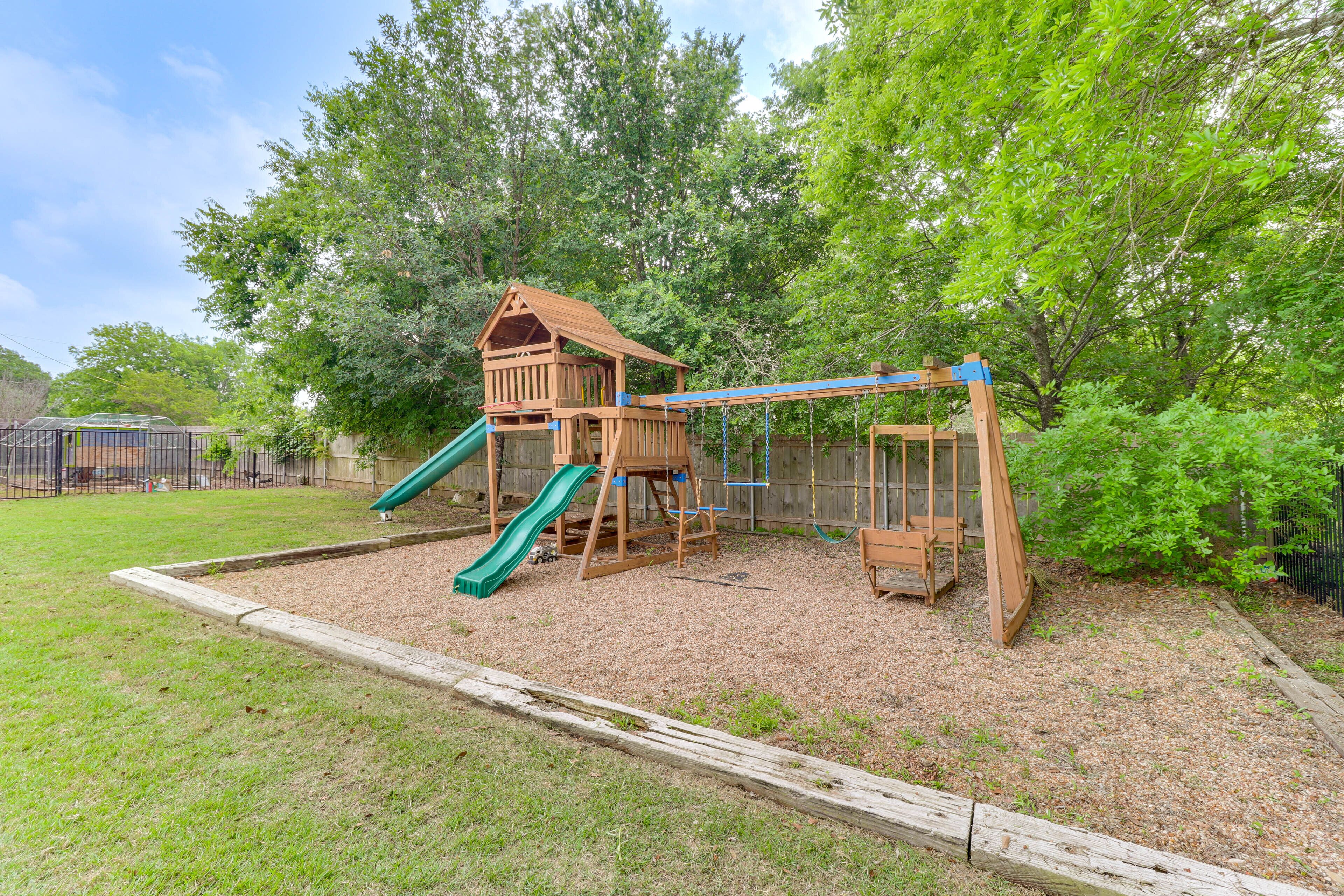 A wooden playground with a slide and swing set, surrounded by a fence and trees.