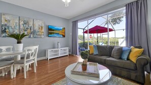 A modern living room with a dining area, a sofa with cushions, a coffee table, and a large window with curtains. - Lake View End Unit Town Home Closest to ALL of the Amenities at WINDSOR HILLS (Kissimmee)