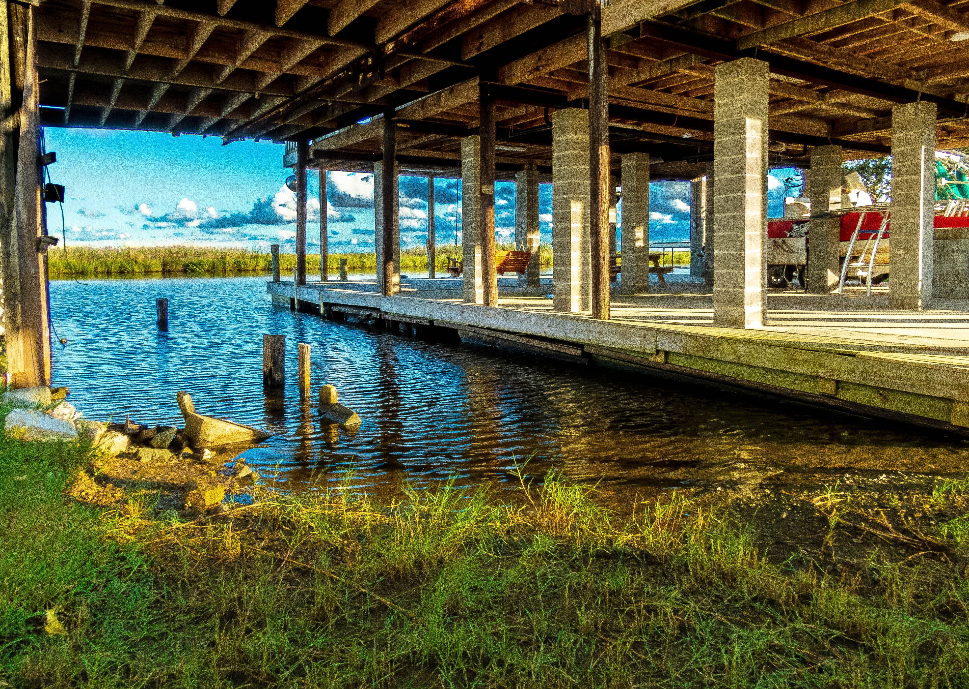 Un muelle que se extiende sobre el agua, sostenido por pilares de concreto, con vistas a un parque y un área de juegos en el fondo.