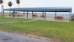 A covered picnic area with benches and picnic tables, a wheelchair-accessible parking sign, and a view of the beach and pier in the background. -  Reel'em Inn - Adventures Start Here! (Port O'Connor)