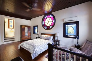 A bedroom with a large bed, a wooden wardrobe, a round stained glass window, and a window with an air conditioning unit. - The Bell Tower Loft at Sanctuary Place (Savannah)