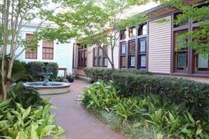 A courtyard with a fountain, surrounded by lush greenery and a building with large windows and a brick pathway.