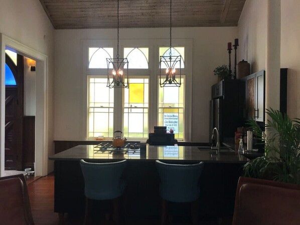 A kitchen with a central island, two hanging pendant lights, and a view through large windows.