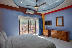 Un dormitorio con cama, cómoda, ventilador de techo, televisión y ventana con vistas a edificios.