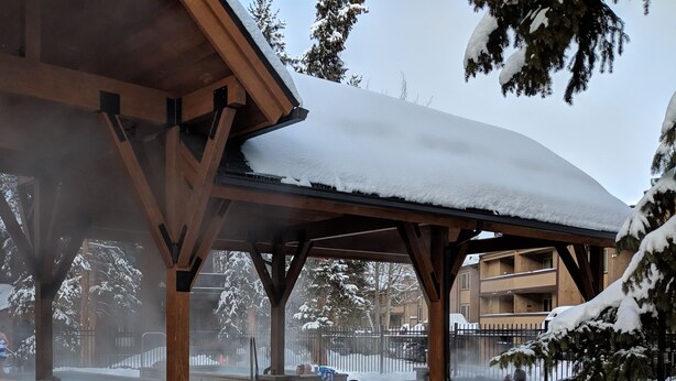 Uma área externa coberta com bancos e uma banheira de hidromassagem, rodeada por árvores e edifícios cobertos de neve.