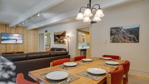 Uma área de refeições com uma mesa de madeira posta para quatro pessoas, rodeada por cadeiras vermelhas. Um sofá de couro marrom e uma televisão são visíveis ao fundo.