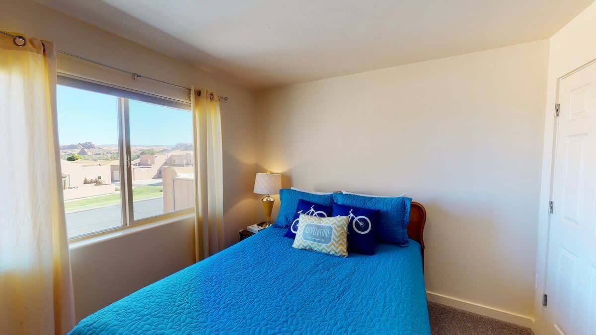 Una camera da letto con un'ampia finestra, un letto con lenzuola blu, un comodino con una lampada e vista sull'esterno.