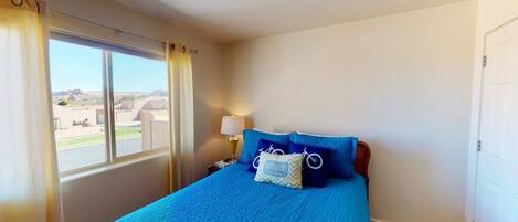 Una camera da letto con un'ampia finestra, un letto con lenzuola blu, un comodino con una lampada e vista sull'esterno.