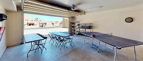 Una habitación con mesa de ping pong, una mesa de comedor con sillas y estanterías con libros.