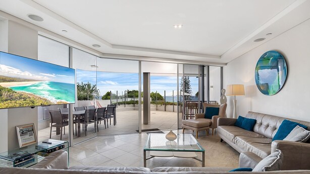 A modern living room with a large TV, comfortable seating, a glass coffee table, and a view of the ocean.