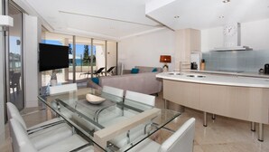 A modern kitchen with a central island, a dining area with glass tables, and a living area with a sofa and TV.