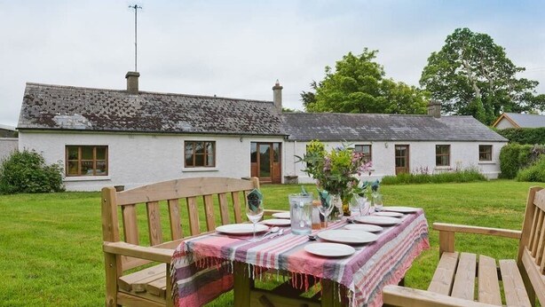 A wooden table set outdoors with a tablecloth, surrounded by a grassy area and a white building with a chimney in the background. - Spacious traditional Irish cottage, on the banks of the Boyne. Pet friendly. (Baltray)