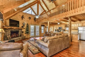 A spacious log cabin interior with a fireplace, a living room with sofas, a kitchen area, and a balcony with a railing.