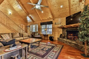 A log cabin interior with a fireplace, a ceiling fan, a staircase, and a Christmas tree.