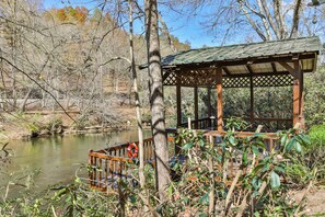 A wooden gazebo with a metal roof overlooking a calm body of water, surrounded by trees and vegetation.