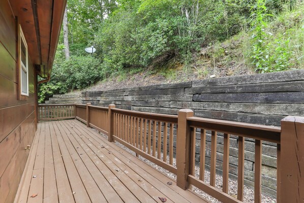 A wooden deck with a railing, surrounded by dense greenery and a stone retaining wall.