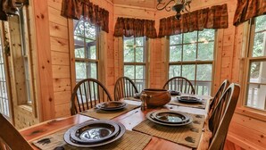 A wooden dining room with a table set for four, surrounded by chairs, and large windows with curtains.