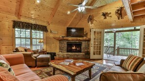 A cozy cabin interior with a fireplace, wooden walls, a ceiling fan, and a deck with a railing.