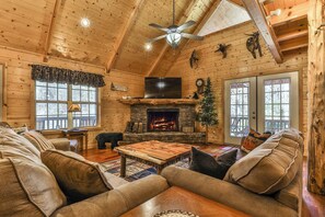 A cozy log cabin interior with a fireplace, leather sofas, a television, and a ceiling fan.