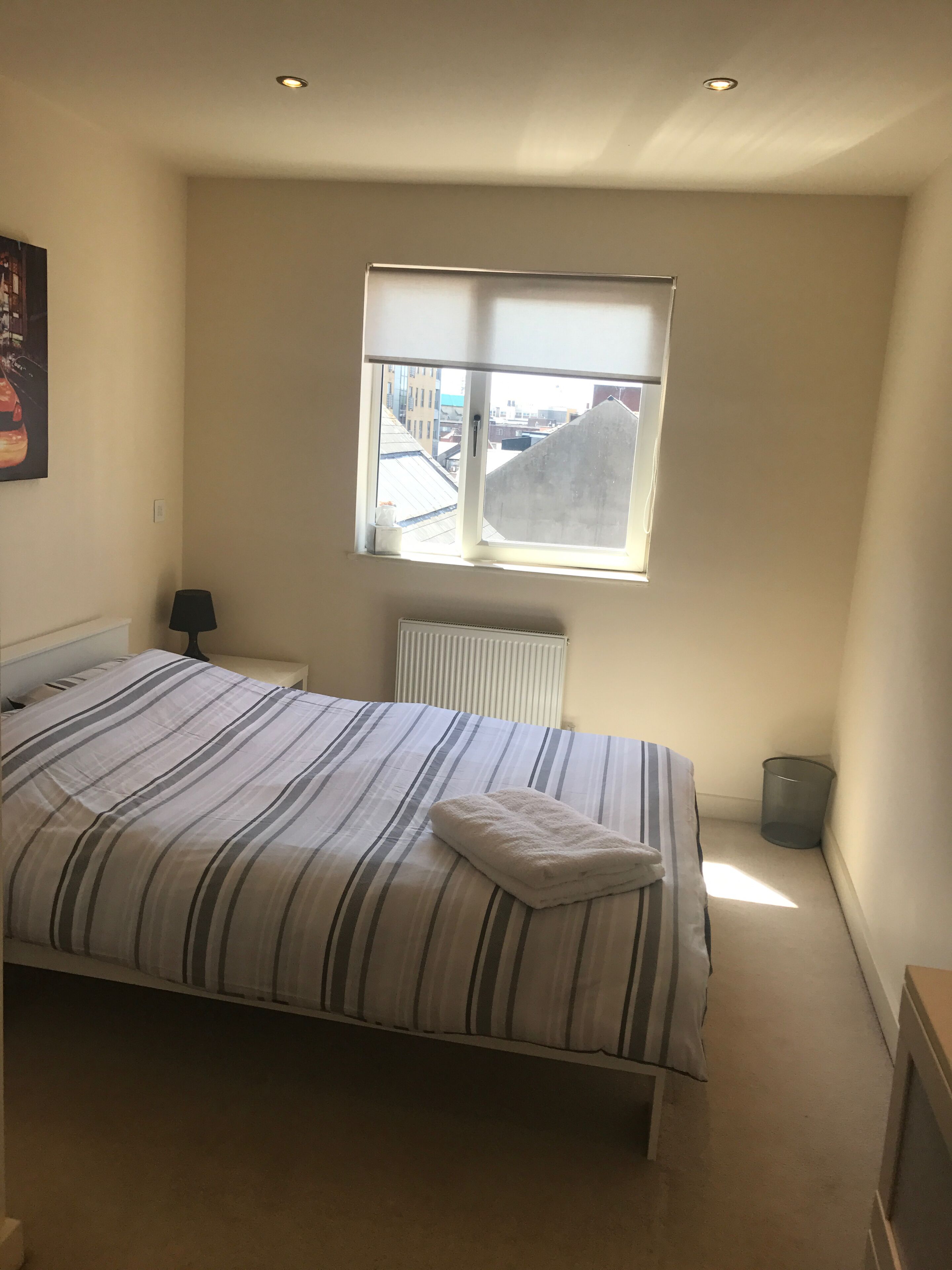 Ein Schlafzimmer mit Bett, Heizkörper, Fenster mit Blick auf Gebäude, Lampe und Mülleimer.