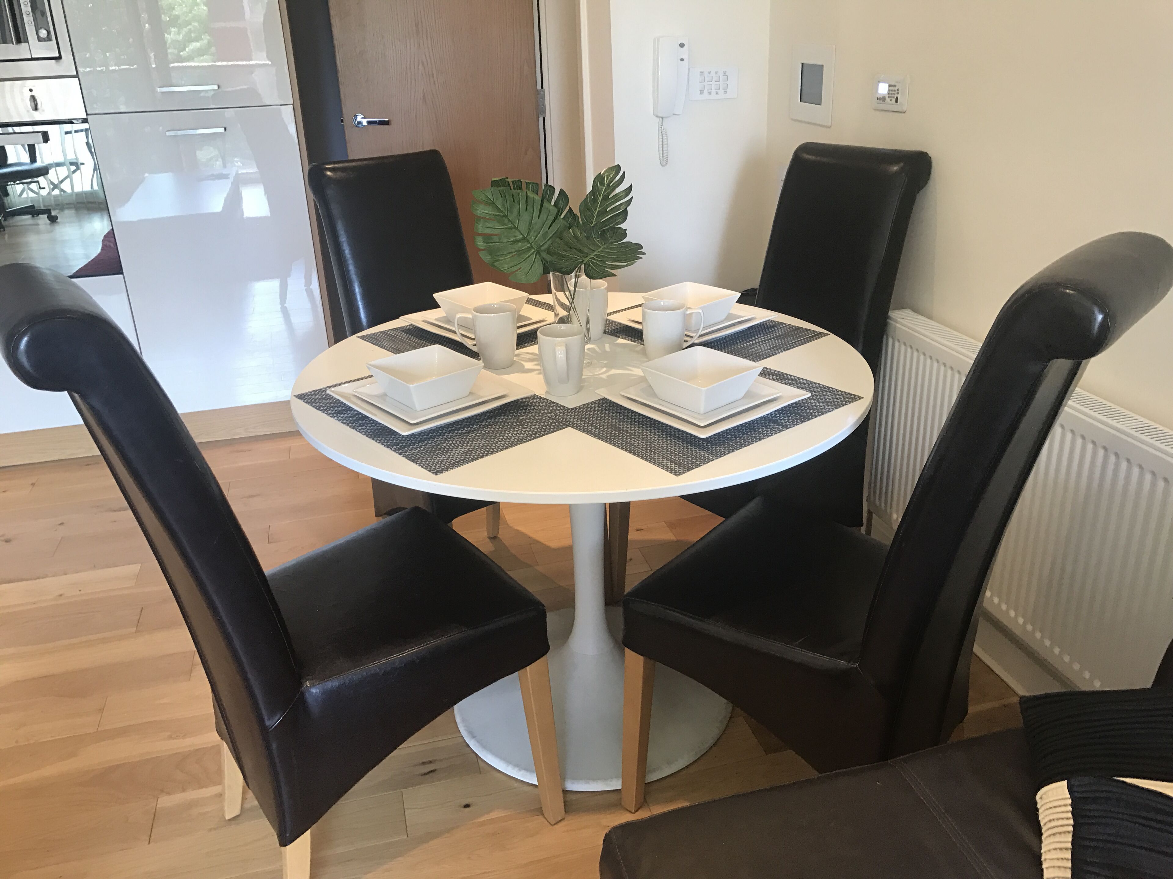 Una mesa redonda de comedor con cuatro sillas negras, platos, tazones y tazas blancos. Hay una planta en maceta sobre la mesa.