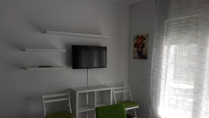 A room with a white table, two green chairs, a white bench, a mounted TV, and a painting on the wall. - Alexis II Zeus Salou 0632 (Salou)