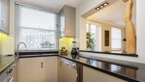 A modern kitchen with a black countertop, stainless steel appliances, and a window with blinds. - Garden Square Serenity, by Veeve (London)