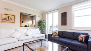 A living room with a white sofa, a dark sofa, a glass coffee table, and a large window. - Garden Square Serenity, by Veeve (London)