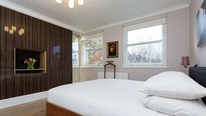 A bedroom with a large bed, a TV mounted on the wall, a window with a view of buildings, and a framed picture on the wall. - Garden Square Serenity, by Veeve (London)