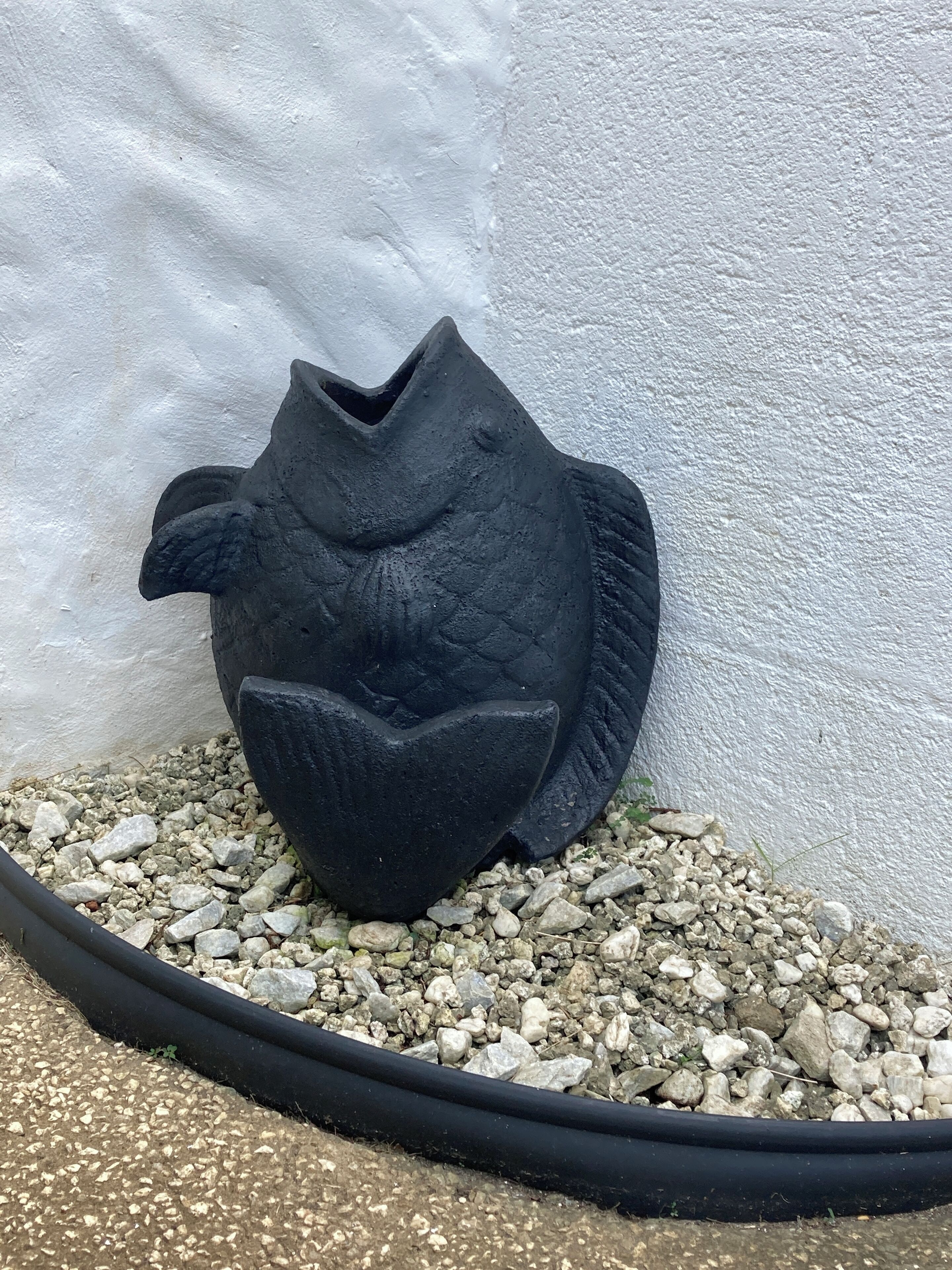 A black, textured sculpture resembling a fish, placed on a bed of small pebbles against a white wall.