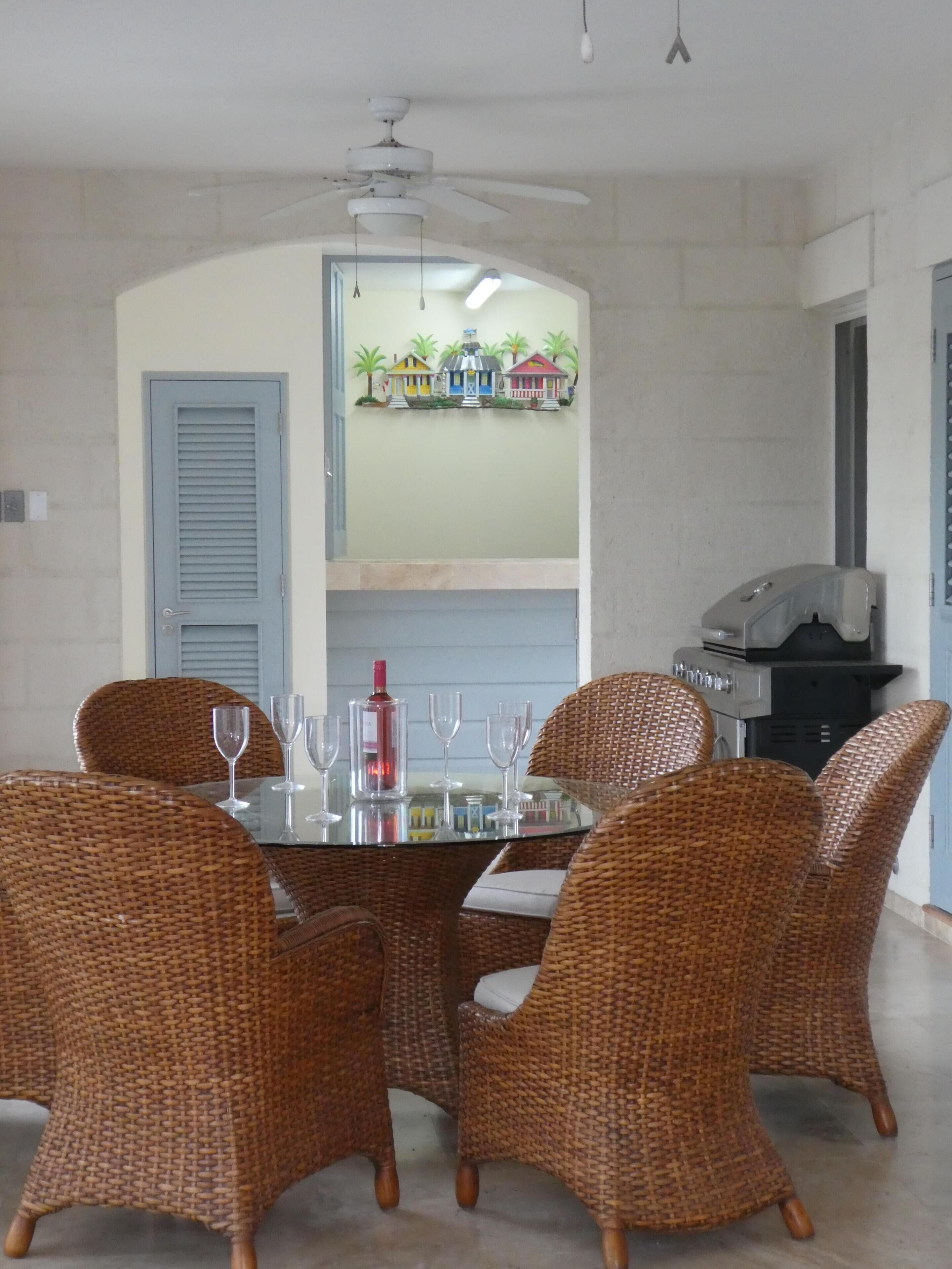 Un coin repas avec des chaises en osier et une table en verre, un ventilateur de plafond et une étagère murale décorée d’objets colorés.