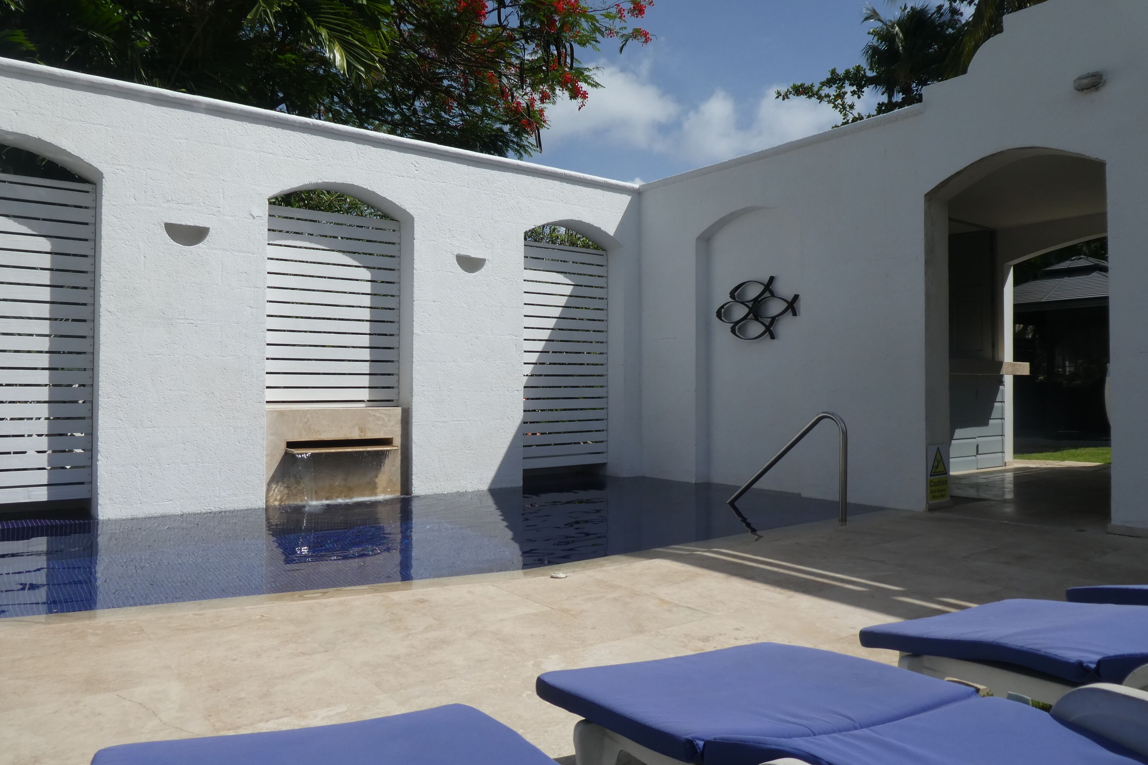 An outdoor swimming pool with lounge chairs, a water feature, and a white wall with arched windows.