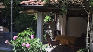 Un área de estar al aire libre techada, con una mesa y sillas, rodeada de macetas con plantas y flores.